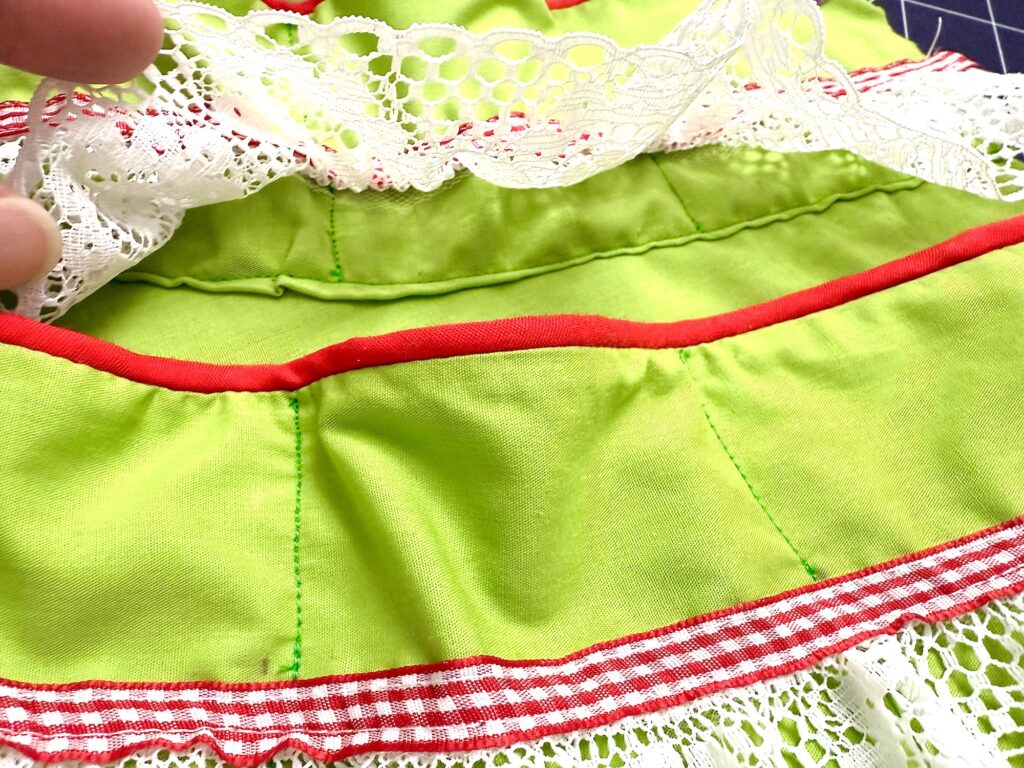 Close-up of an advent tree made from green fabric with red stitching, accented by a red and white gingham ribbon and delicate white lace trim. A hand lifts part of the lace for a detailed view.