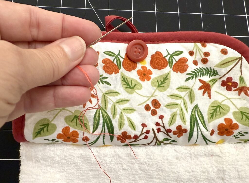 A hand holds a needle with orange thread, sewing a red button onto a fabric piece with a floral pattern, above a white towel on a black grid surface.