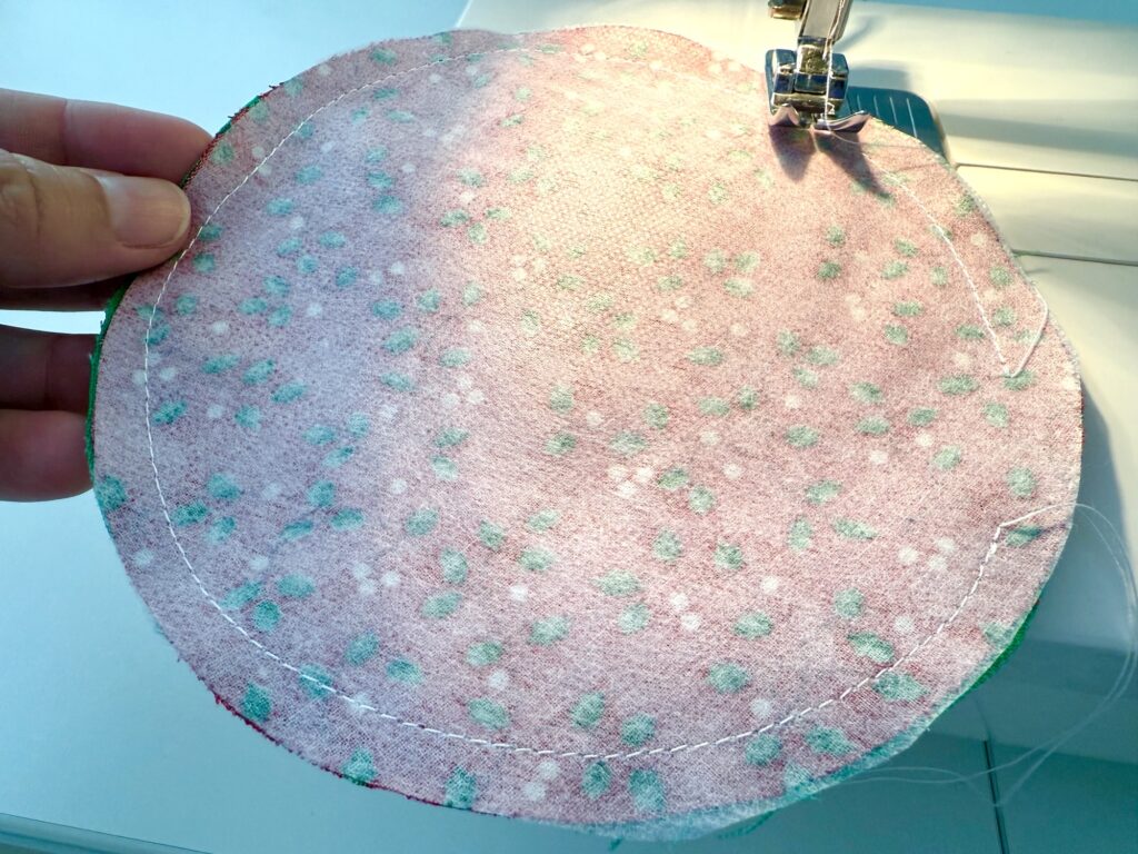 A close-up of a person's hand holding a round piece of fabric with green and pink dots, being sewn with a sewing machine along the edge.