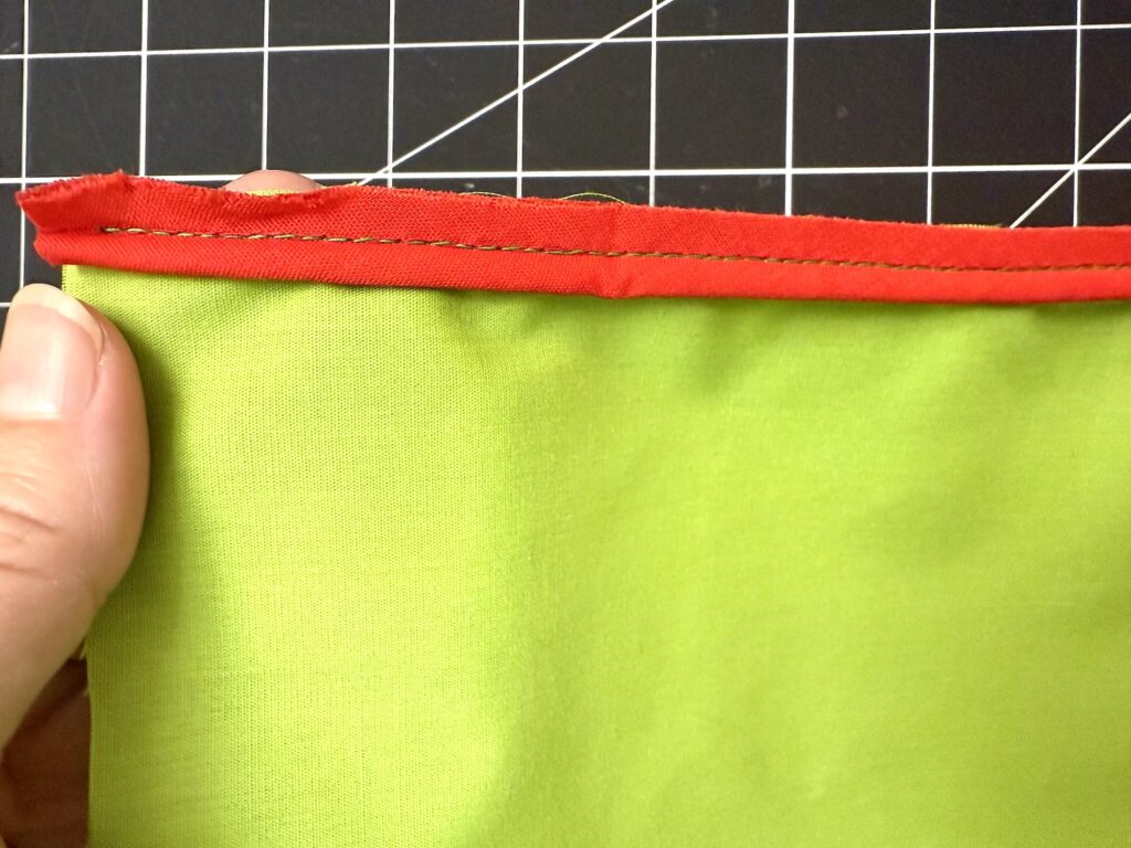 A hand holds green fabric with red bias tape sewn along the edge—an ideal start to an advent tree project; white stitching stands out, and a grid cutting mat appears in the background.