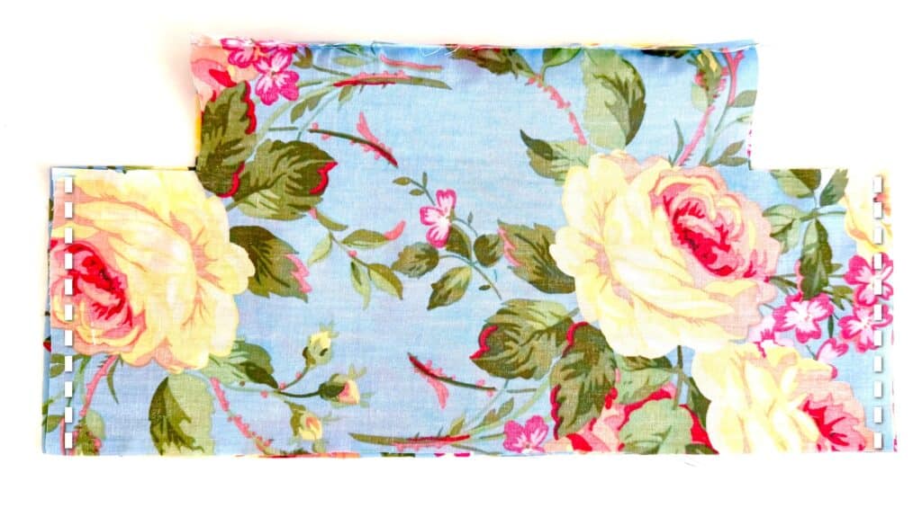 A piece of blue floral fabric with yellow and pink flowers, cut in a rectangular shape with corners removed and edges pinned, perfect for creating a charming tissue box cover.