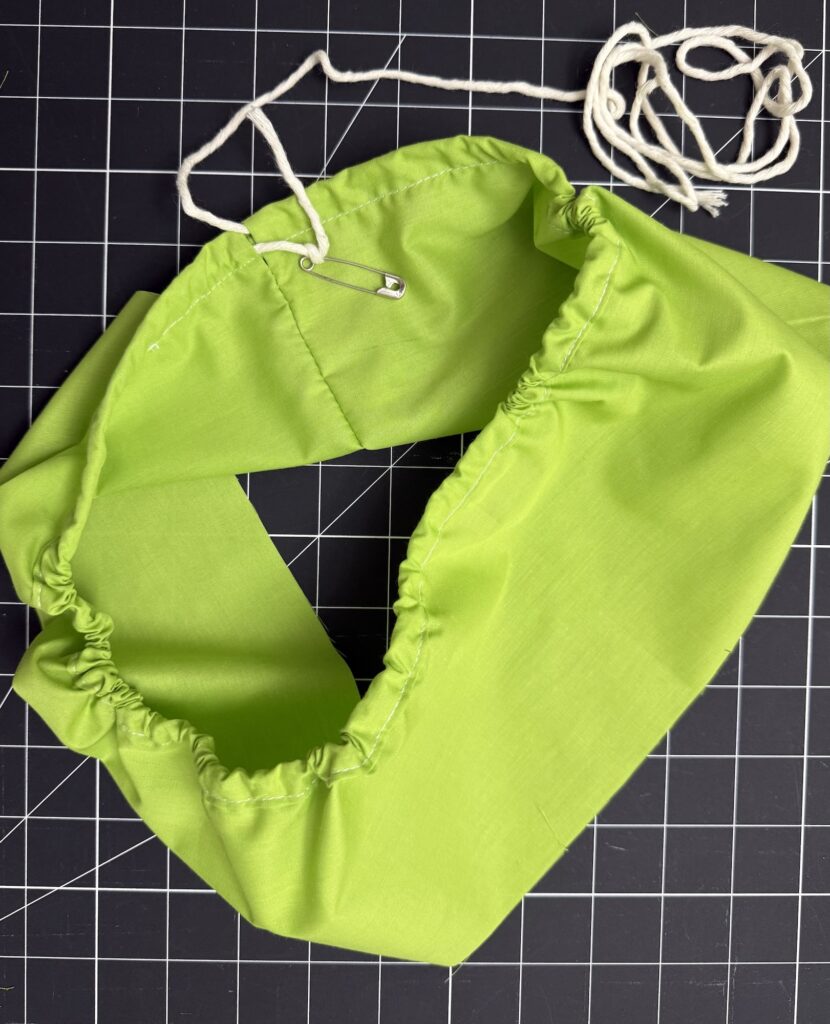 A partially sewn green fabric tube, perfect for an advent tree project, with a drawstring and safety pin on a black grid cutting mat.
