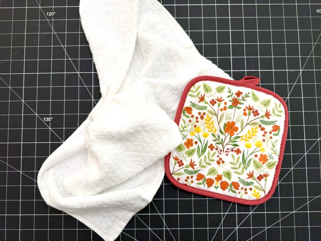 A white kitchen towel and a floral-patterned potholder with a red border are placed on a black grid cutting mat.