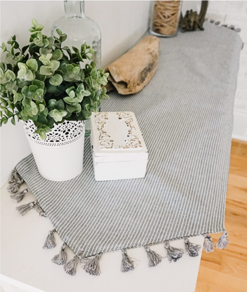 A potted plant, a decorative white box, and a glass bottle sit on a striped table runner with tassels, placed on a light-colored surface.