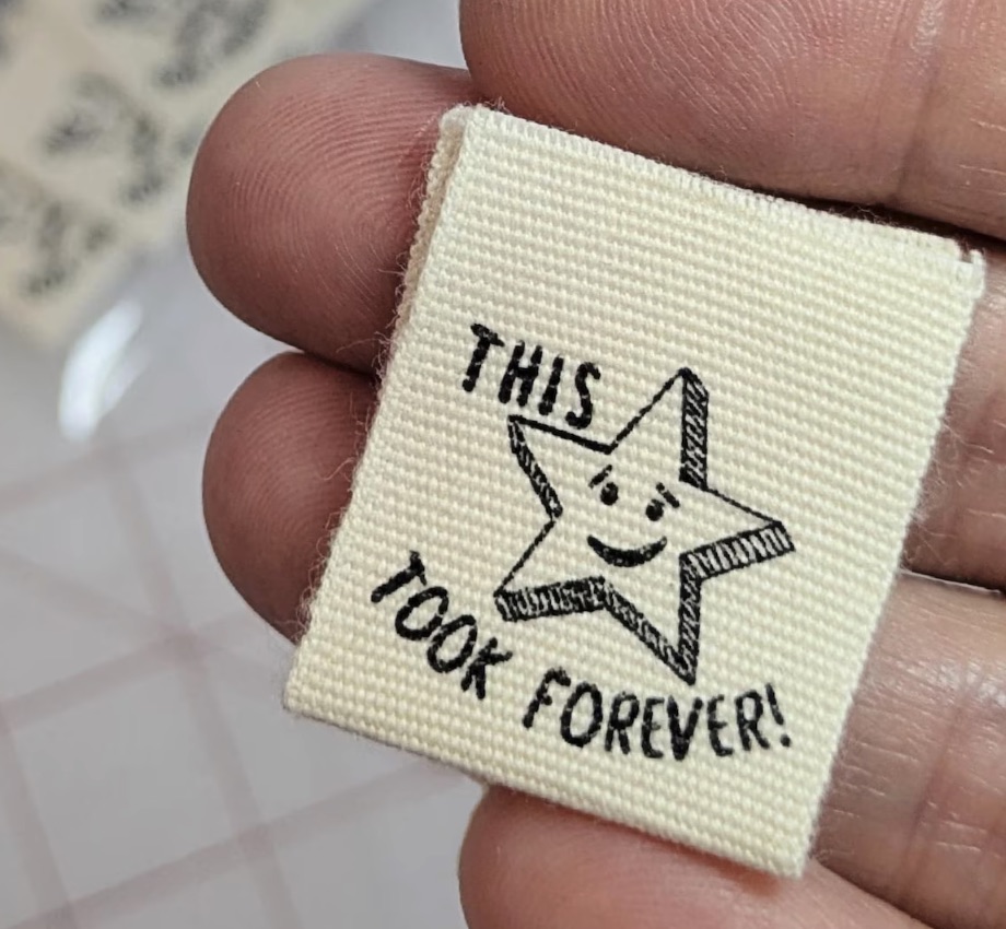hand holding a cream colored sewing label with a star and the text "this took forever"