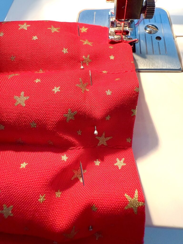 A red fabric with gold stars, pinned in pleats, is being stitched with a sewing machine.