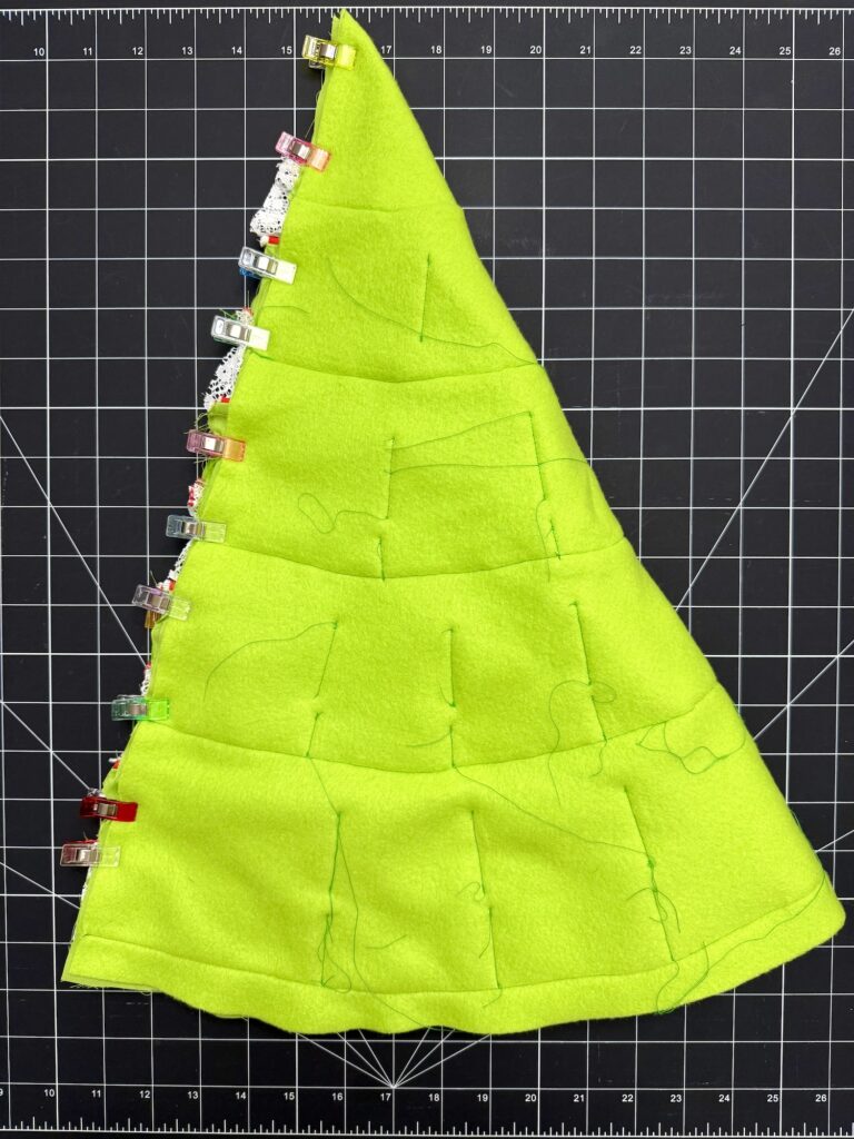 A piece of bright green fabric, perfect for an advent tree, cut and sewn into a triangle shape with sewing clips along one edge, placed on a black grid cutting mat.