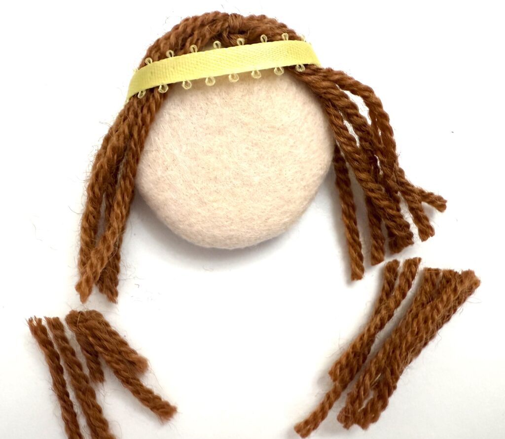 A round beige object with brown yarn strands attached as hair and a yellow band on top; several yarn strands are cut and placed below.