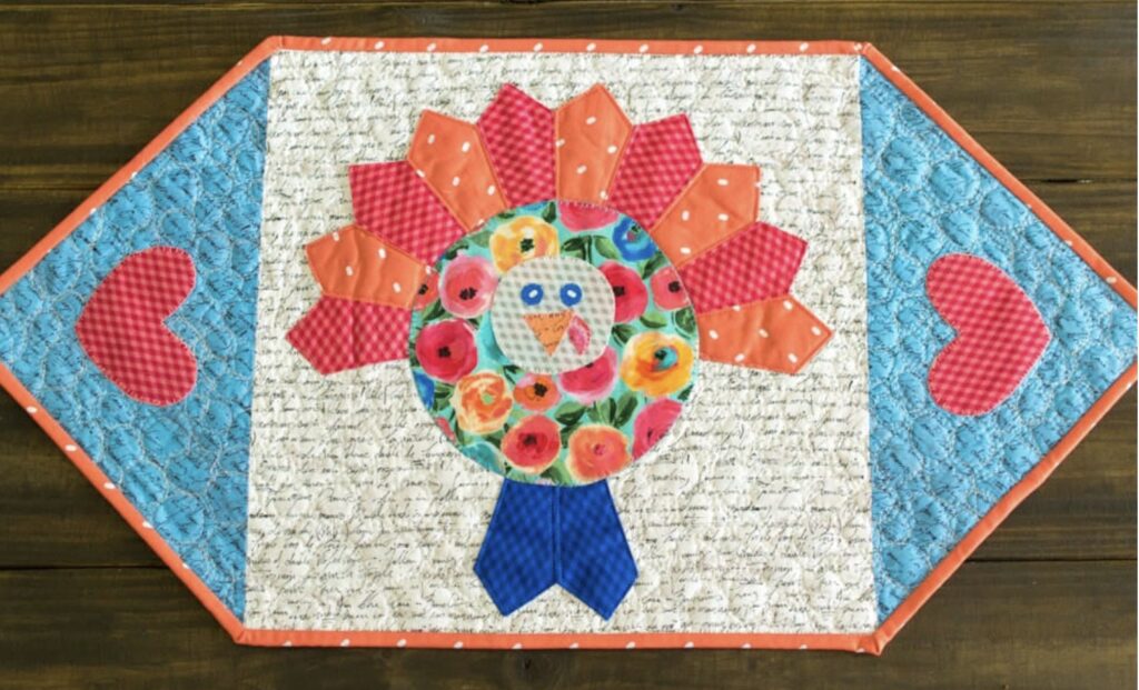 A quilted table runner featuring a turkey with a floral body, red checkered feathers, and blue checkered wings, bordered by blue diamond-shaped ends with pink hearts.
