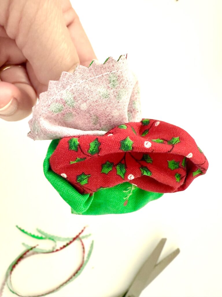 A hand holds a small bundle made of red, green, and pink patterned fabric; scissors and a piece of ribbon lie on a white surface below.