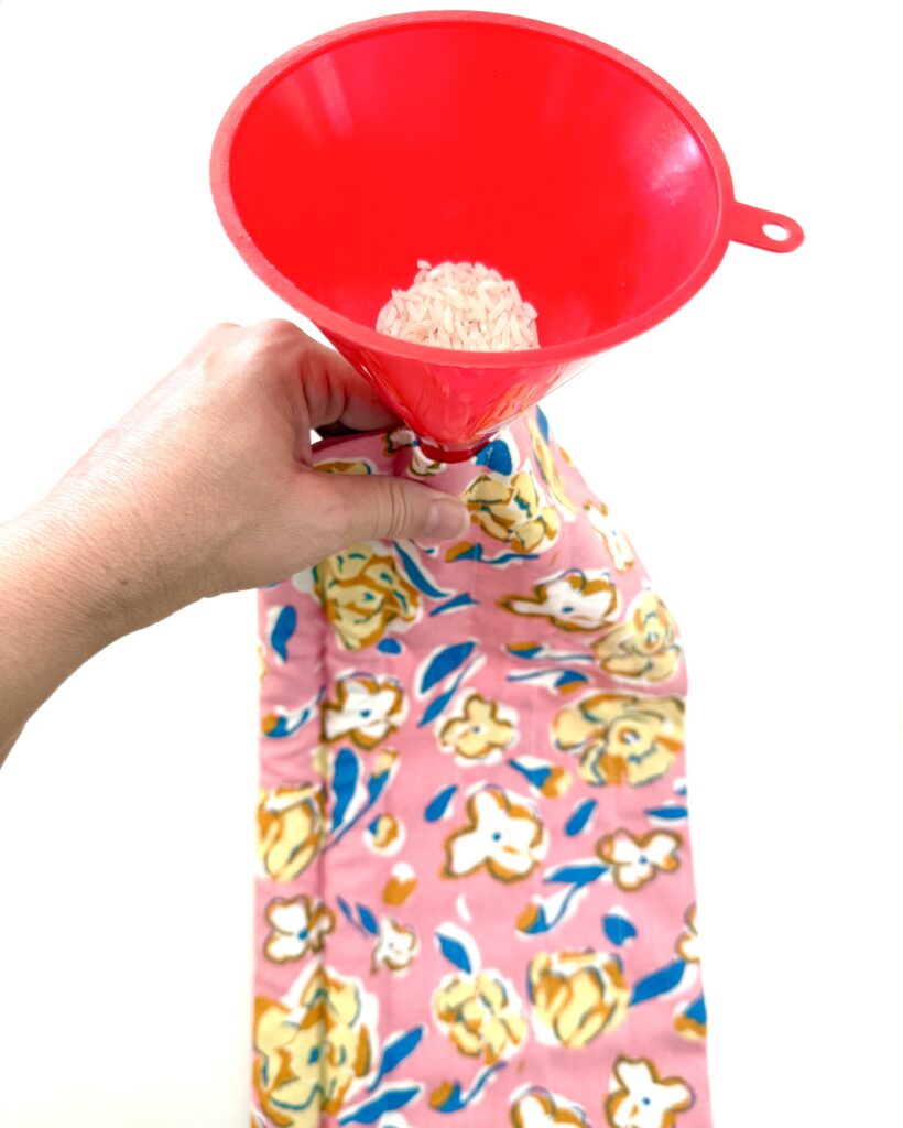A hand holds a pink floral fabric trivet while pouring rice through a red funnel into the fabric against a white background.