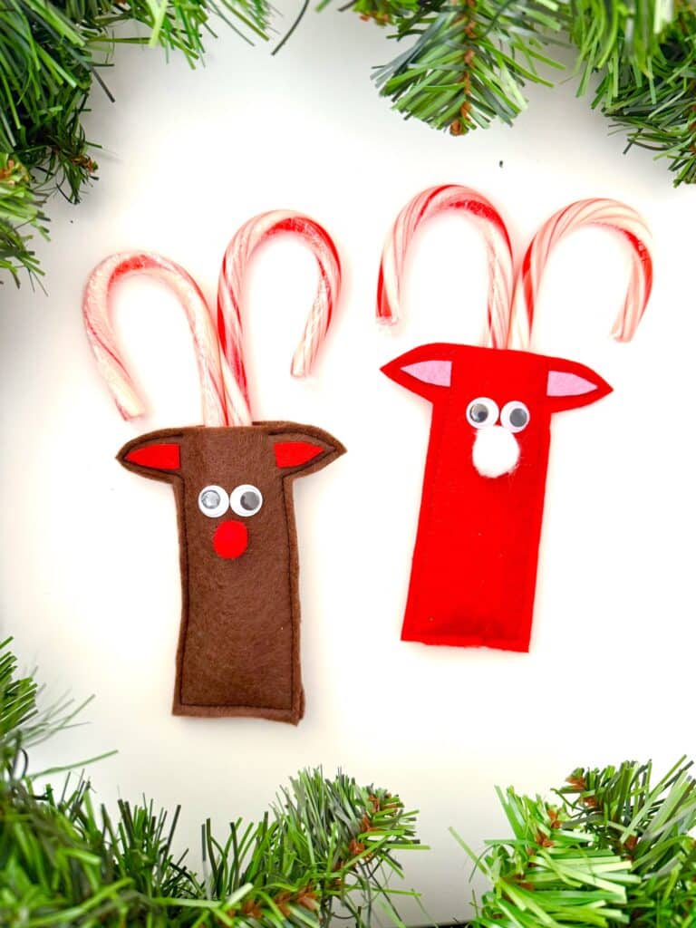 Two candy canes are decorated with felt to resemble reindeer, one in brown and one in red, each with googly eyes and a nose, surrounded by green garland.