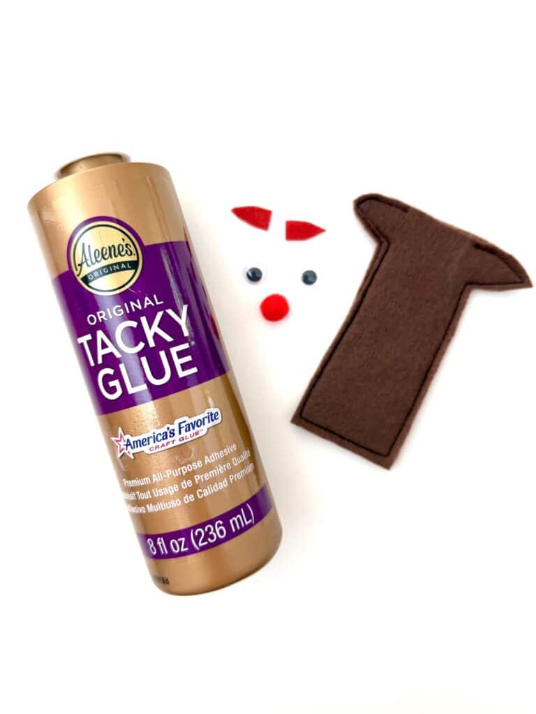 A bottle of Aleene’s Original Tacky Glue, two googly eyes, red felt shapes, and a brown felt piece shaped like a reindeer head are arranged on a white background.