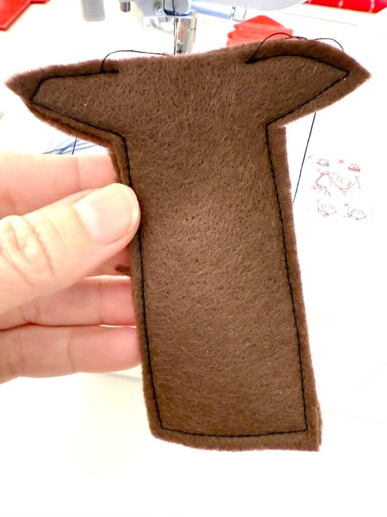 A hand holds a brown piece of felt shaped like a capital "T" with black stitching around the edges; a sewing machine is visible in the background.