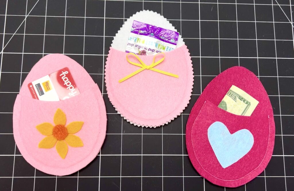 Three felt egg-shaped pouches, each with a pocket holding items like gift cards, candy, cash, or even a small seed packet, are displayed on a cutting mat.