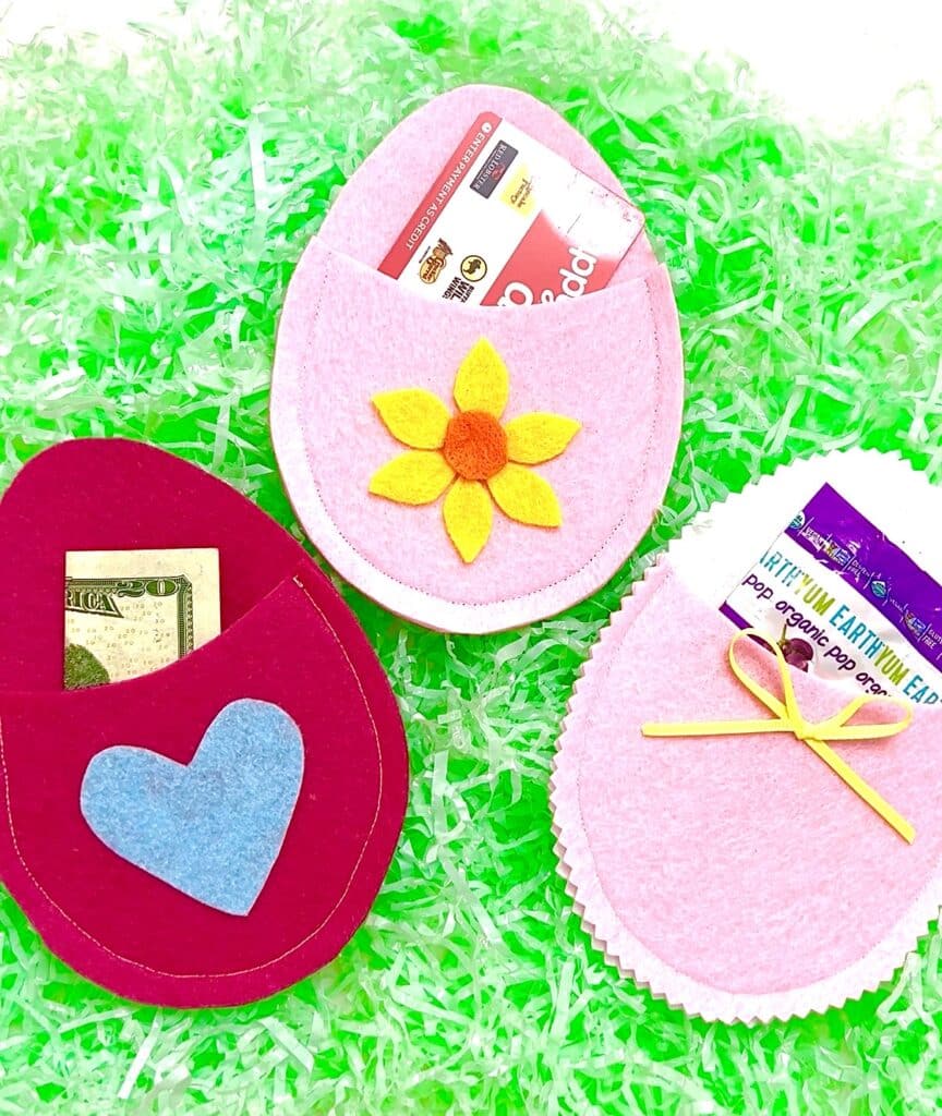 Three felt egg-shaped pouches in pink and red, decorated with a flower, heart, and bow, each holding a gift card, cash, or seed packet, placed on green Easter grass.