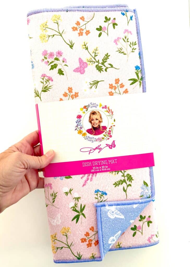 A hand holds a floral and butterfly patterned dish drying mat wrapped in packaging featuring a woman's portrait and the name "Dolly.