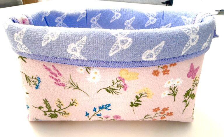 A rectangular fabric basket made from a dish mat with a light pink floral pattern on the outside and a folded lavender rim with white butterfly designs.