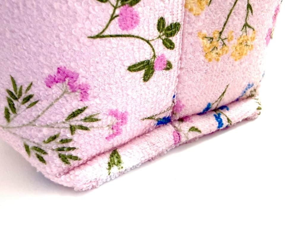 Close-up of a pink textured fabric with embroidered floral designs in green, yellow, purple, and blue, showing a folded edge detail.