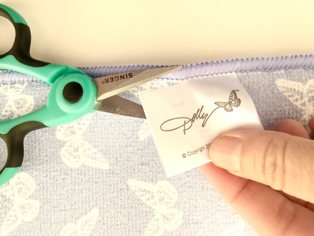 A hand holds a fabric label with "Polly" and a butterfly logo, while green-handled scissors cut the label from a light purple, butterfly-patterned cloth.