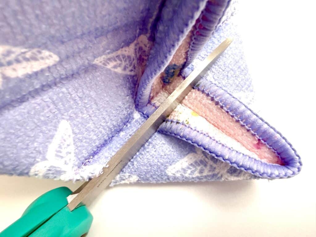 Close-up of scissors with a green handle cutting along the seam of a folded piece of textured, purple fabric with white patterns.