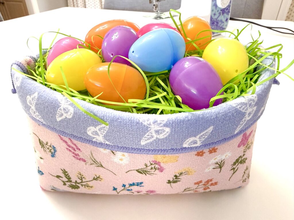 A fabric basket decorated with flowers holds green Easter grass and several colorful plastic Easter eggs.