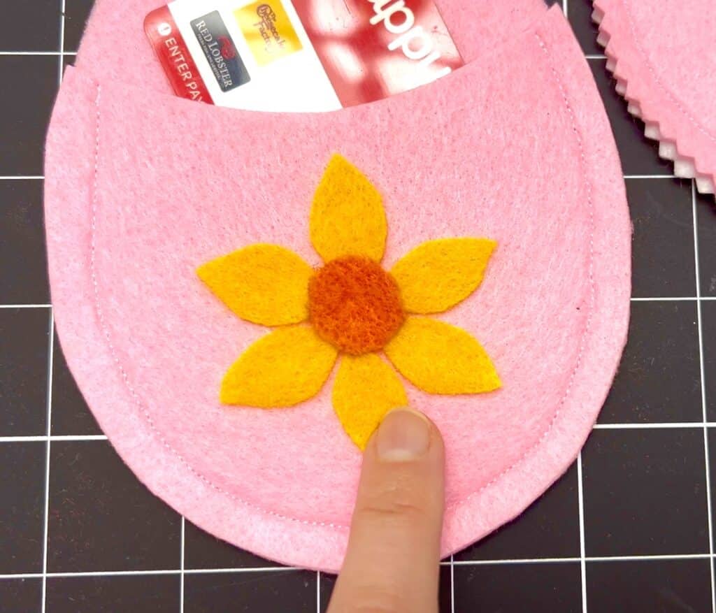 A hand points to a yellow and orange felt flower sewn onto a pink felt slipper, with a partially visible seed packet card inserted inside.