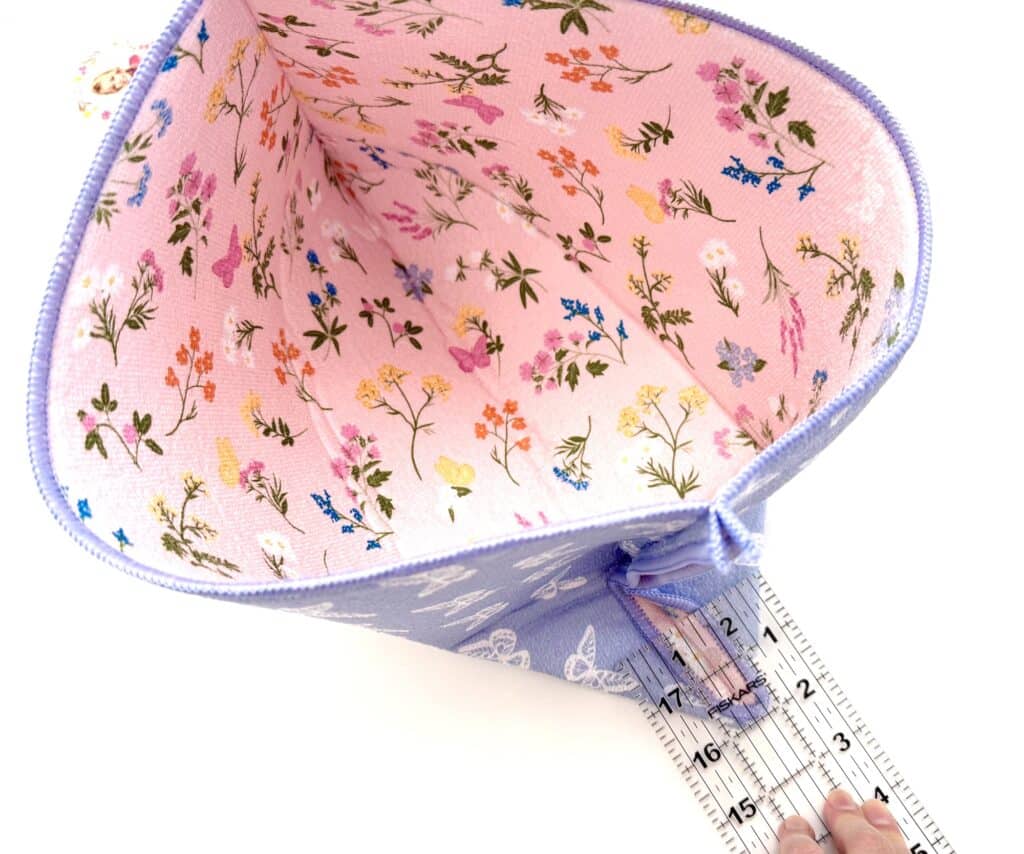 A hand holds a ruler inside a pink, floral-patterned fabric pouch with a purple zipper, measuring the interior width.