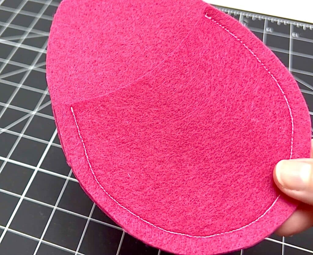 A hand holding a piece of bright pink felt with visible white stitching along the curved edge, resembling a seed packet, placed on a black grid cutting mat.