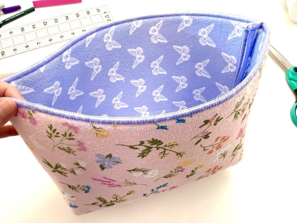 A hand holds an open, rectangular fabric pouch with a floral exterior and a light blue interior patterned with white triquetra symbols. Scissors and a ruler are nearby on a white surface.