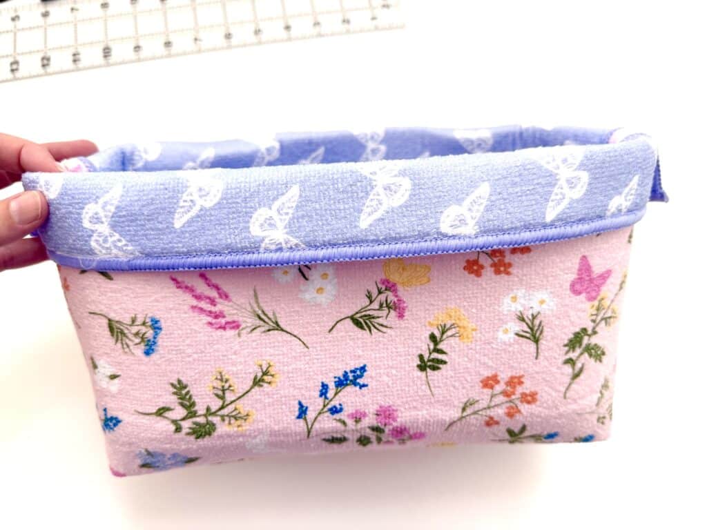 A hand holds a small fabric basket with a pink floral pattern and a blue rim, set on a white surface with part of a clear ruler in the background.