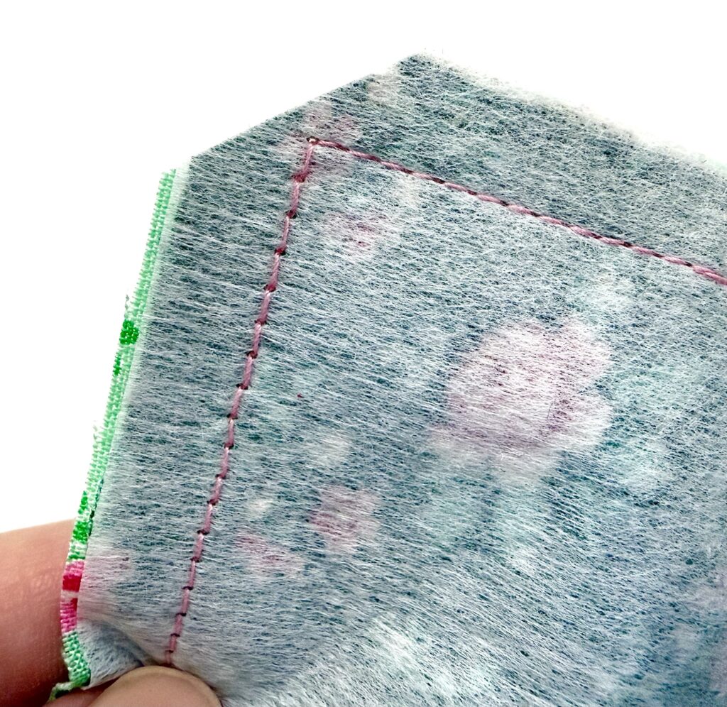 Close-up of a person's hand holding two layers of fabric with visible pink machine stitching along the edges, crafting a charming tea wallet.