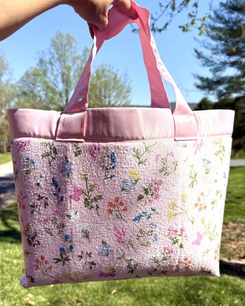 A hand holds a pink tote bag with floral embroidery and pink handles, outdoors on a sunny day with trees in the background.