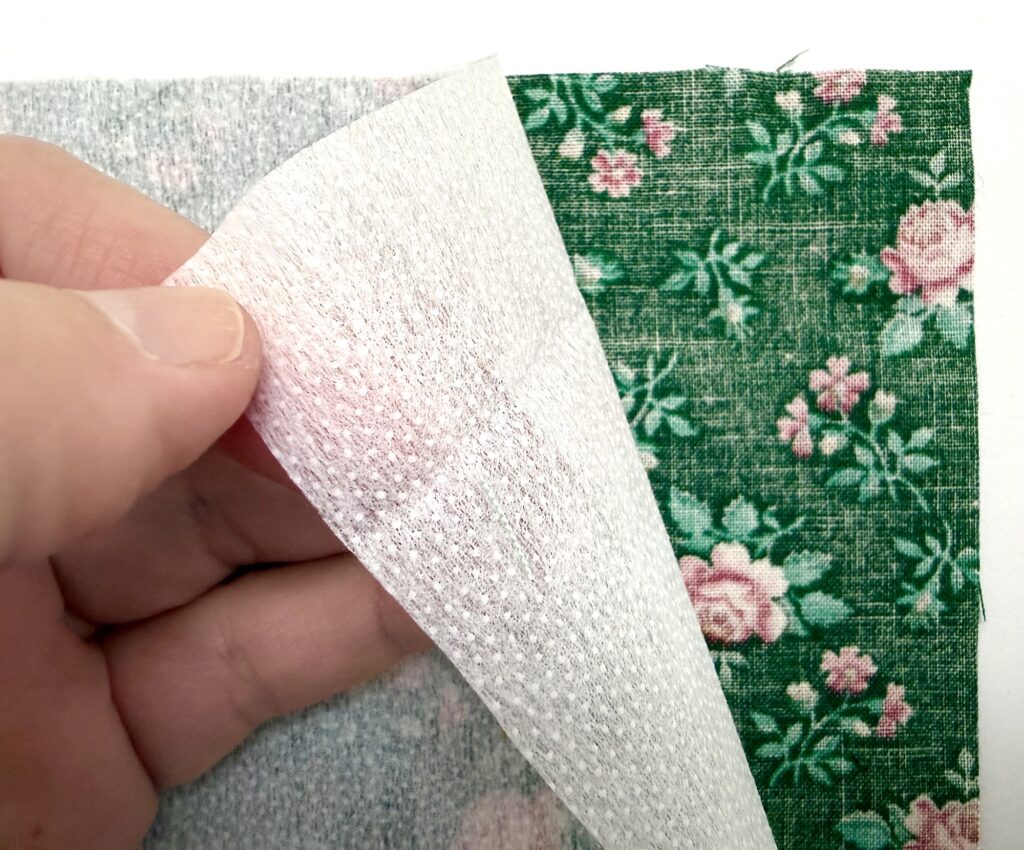 A hand lifts a white fusible interfacing from the back of a green floral fabric, showing the two layers separated—perfect for crafting projects like a tea wallet.