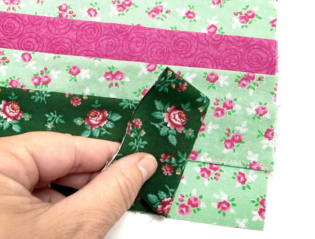 A hand holds a strip of dark green floral fabric over pink and green floral strips, likely in the process of quilting or sewing a tea wallet.