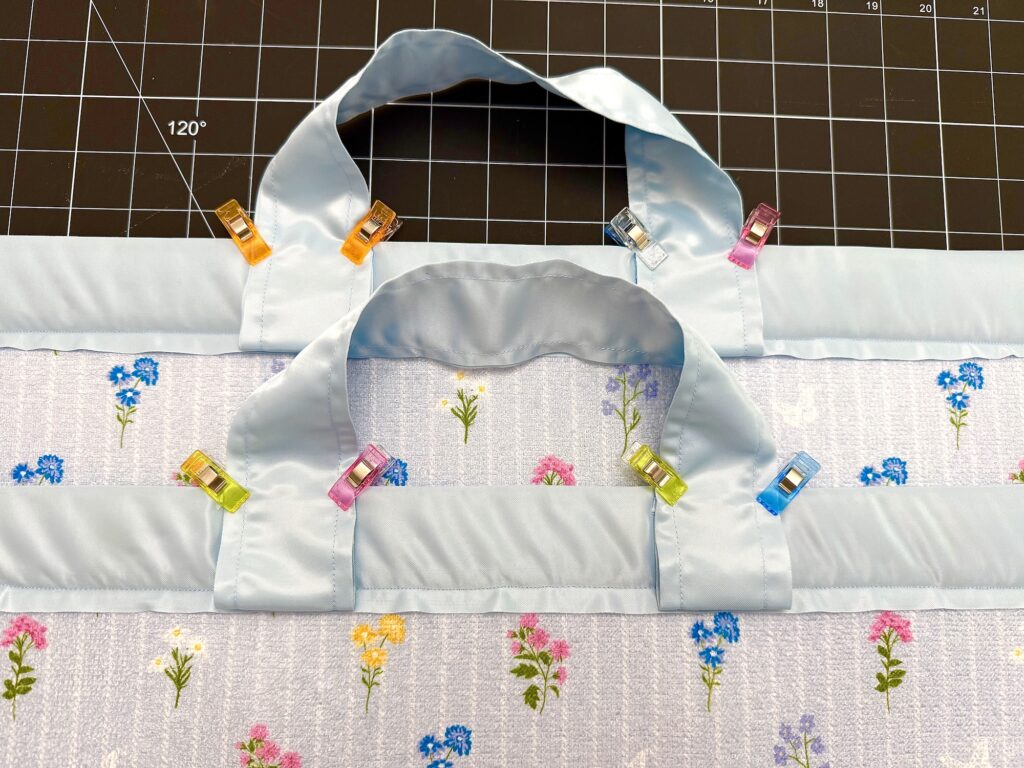 Two pieces of floral fabric with blue borders, perfect for a dish mat or tote bag, are clipped together with colorful sewing clips, set on a cutting mat marked with grid lines and angles.