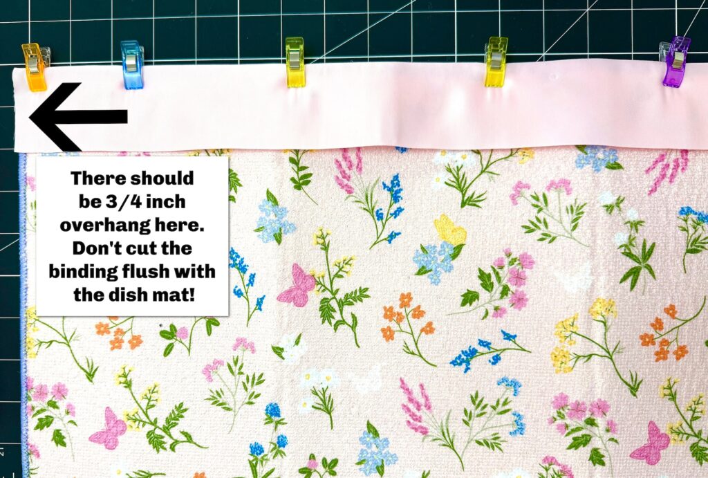 A piece of floral fabric for a dish mat or tote bag is topped with a white binding strip, clipped in place, with a &frac34; inch overhang indicated by an arrow and instructional text.