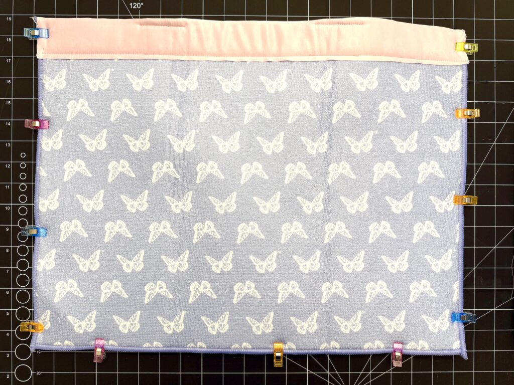 A rectangular piece of fabric with a white butterfly pattern and a pink strip at the top is clipped along the edges with multicolored sewing clips on a gridded cutting mat.