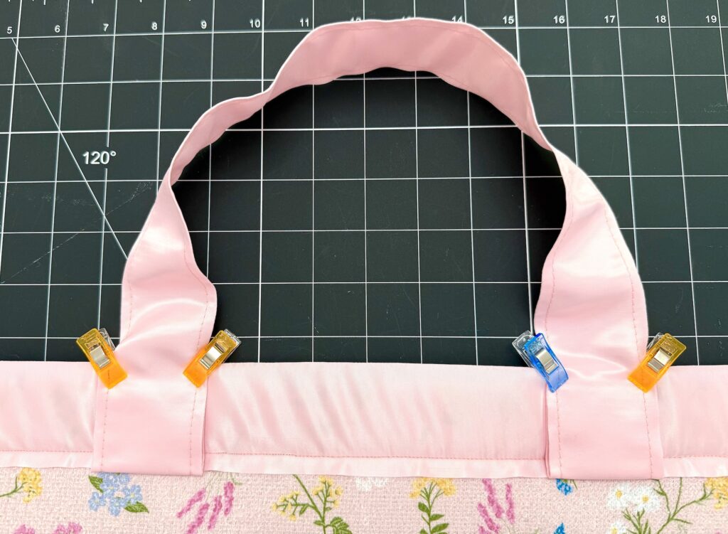 A pink fabric handle is clipped to the top edge of a floral-patterned fabric piece using colored sewing clips, shown on a black cutting mat with a grid.