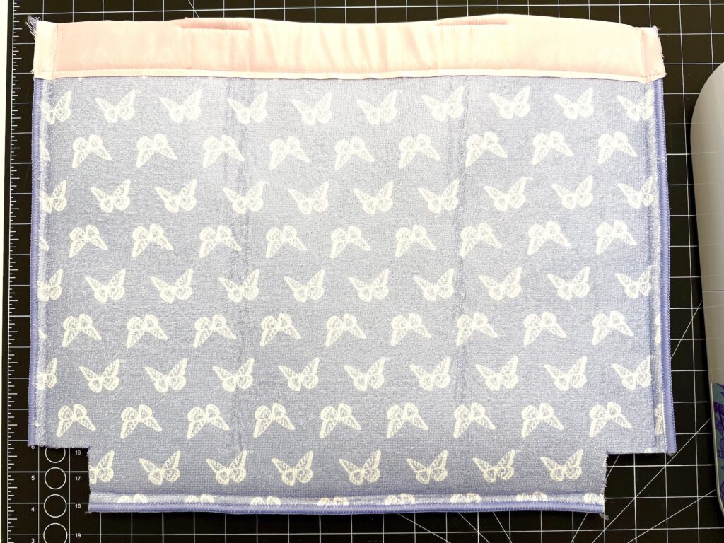 A piece of light blue fabric with white butterfly patterns and a pink strip along the top edge is laid flat on a gridded cutting mat.