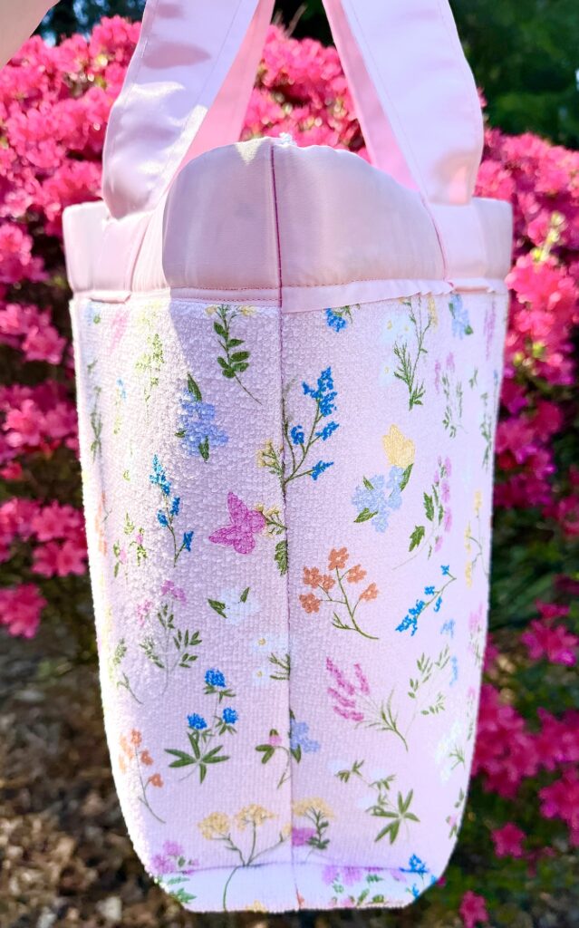A pink dish mat tote bag with floral and butterfly patterns is held outdoors in front of bright pink flowering bushes.