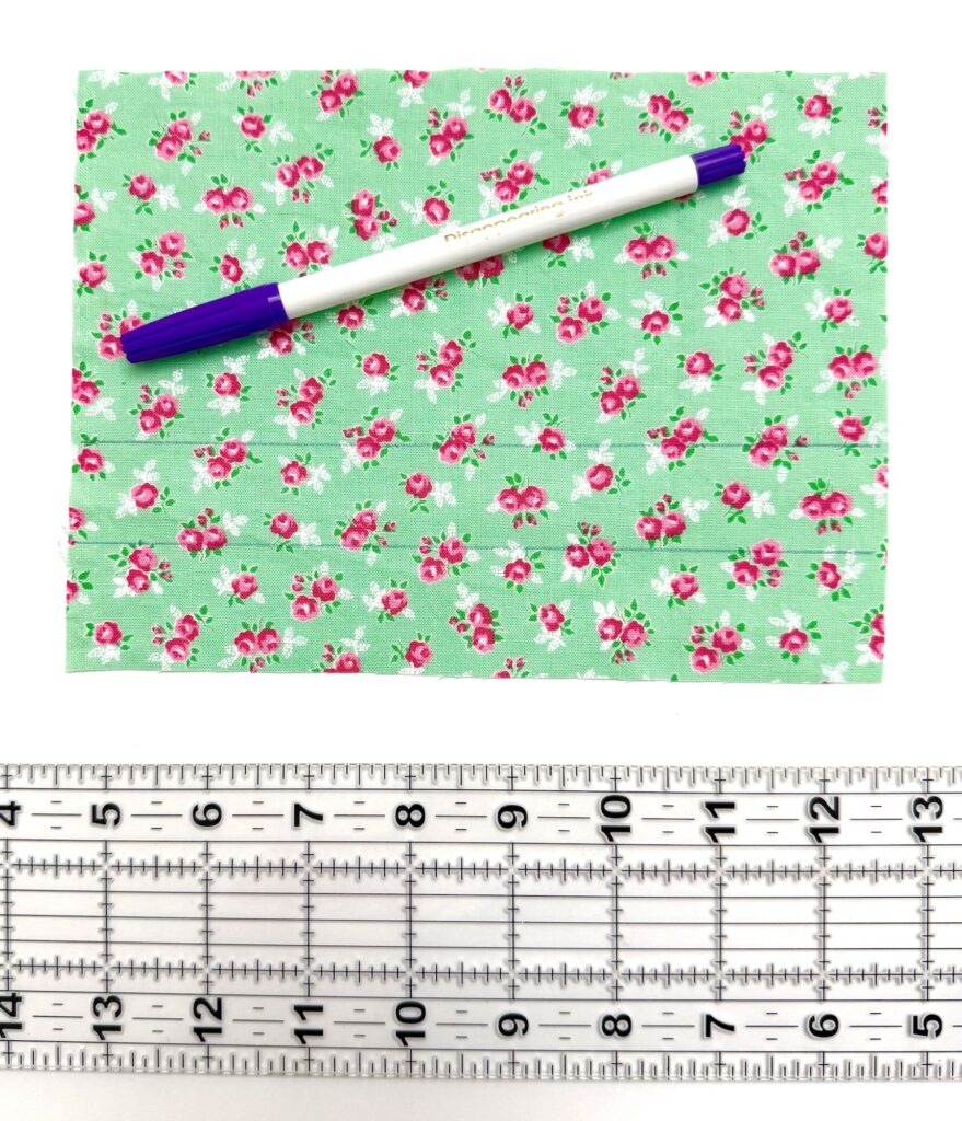A blue and white pen rests on green floral fabric, possibly part of a tea wallet, placed above a clear plastic ruler with black markings on a white background.