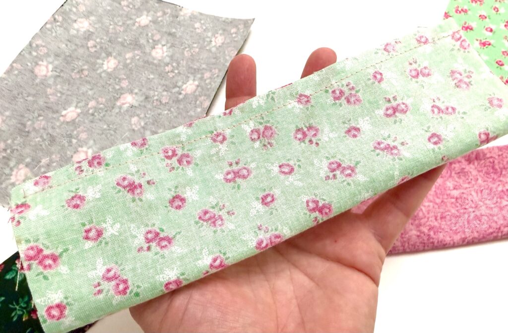 A hand holds a piece of light green fabric with a pink floral pattern, perfect for crafting a unique tea wallet; more fabric pieces with different patterns are in the background.