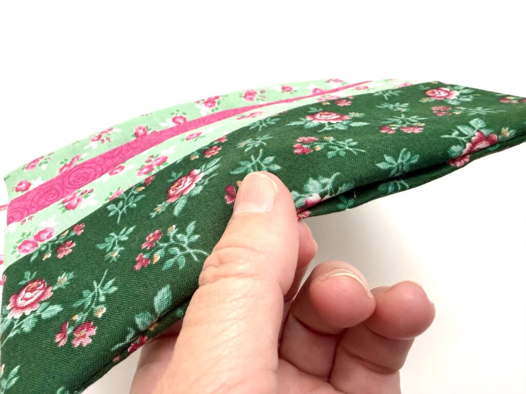 A hand holds a tea wallet made of soft, padded fabric with green, pink, and floral patterned strips against a white background.