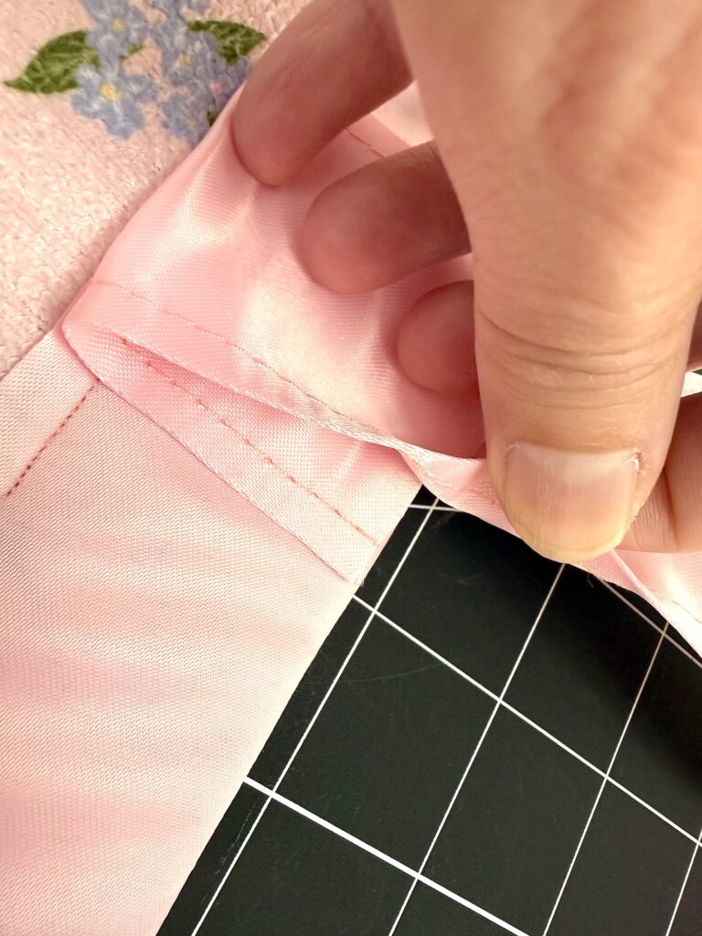 A close-up of a hand folding the corner of pink satin fabric with visible stitching, positioned over a black and white grid cutting mat—perfect detail for a dish mat or tote bag project.