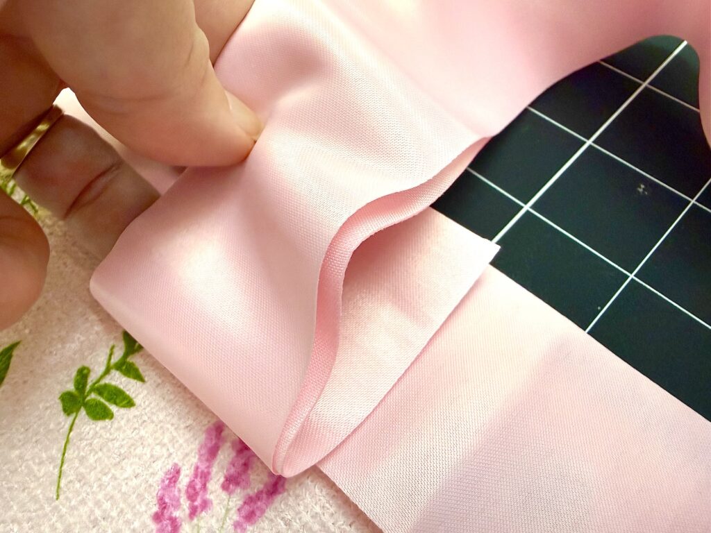 A hand holds the edge of light pink fabric with a folded hem, positioned on a cutting mat with a grid pattern.