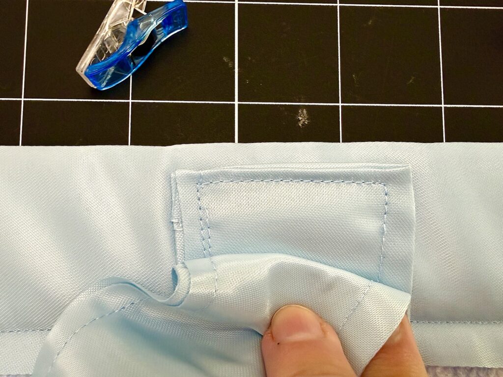 A hand holds light blue fabric with a sewn square patch, ideal for a dish mat or tote bag; a sewing clip and black grid cutting mat are visible in the background.