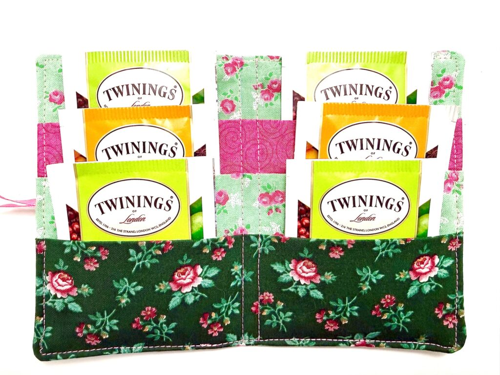 A floral fabric tea wallet holds six Twinings tea packets in green, yellow, and orange packaging, displayed upright in two rows.