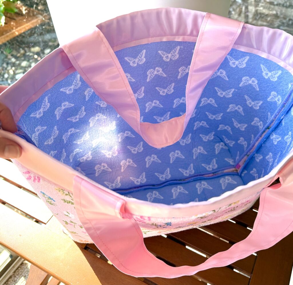 A hand holds open a pink tote bag with a blue butterfly-patterned lining, shown on a wooden table in sunlight.