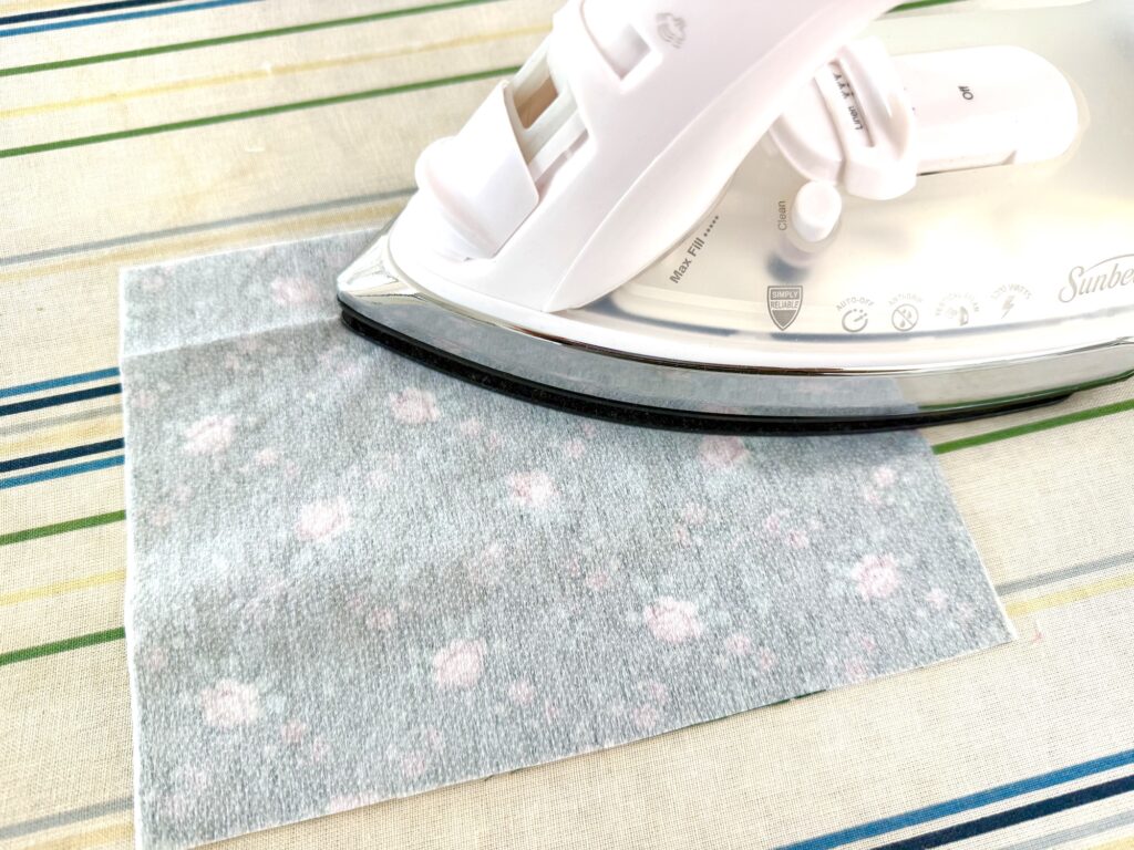 A white steam iron presses a piece of light gray floral fabric, perfect for crafting a charming tea wallet, on a striped ironing board.
